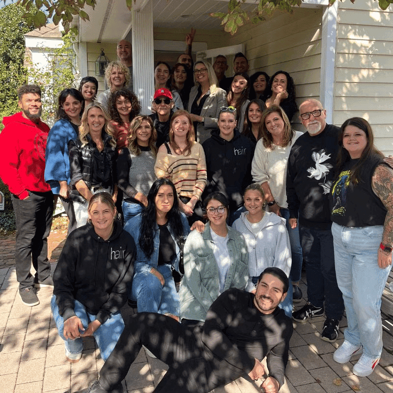 Group photo of people smiling outside a house under tree shade.